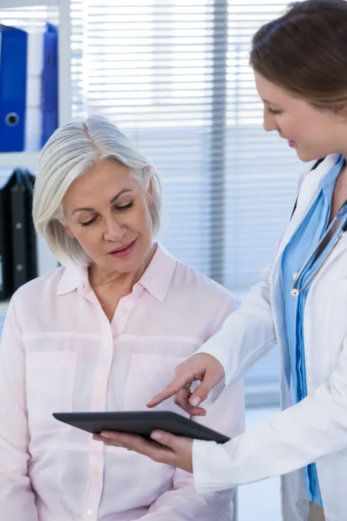 A female doctor in a white coat hold a tablet and talks to an older female patient in a light pink shirt.